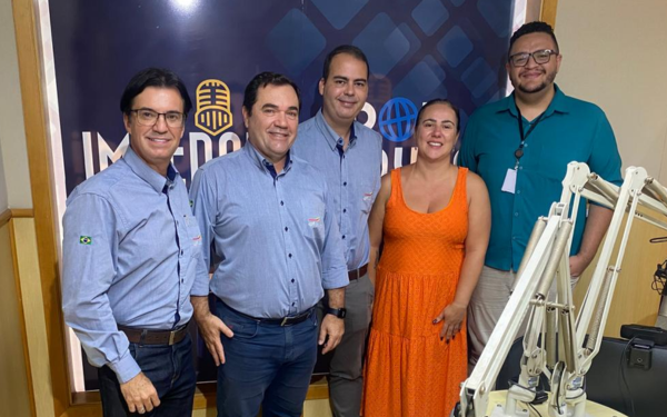 Cinco pessoas estão em pé, lado a lado, dentro de um estúdio de rádio. Ao fundo, há uma parede com a identidade visual da Rádio Zebu FM. À esquerda, Alexandre Alonso, de camisa azul clara e óculos, sorri. Ao lado dele, Clauber Pazin, também de camisa azul clara, sorri com as mãos cruzadas. Ao centro está Gabriel Oliveira, com camisa parecida e um leve sorriso. À direita dele está Jacqueline Galgani, usando um vestido laranja vibrante, sorridente. Na ponta direita está Eudis Filho, de camisa verde e crachá no pescoço, também sorrindo. Em primeiro plano, aparecem braços articulados de microfones do estúdio de rádio.