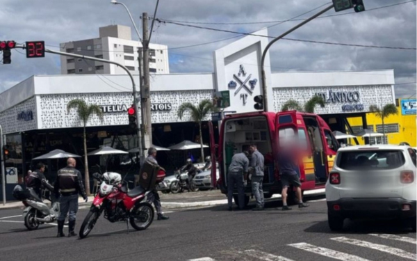 A imagem mostra uma cena de atendimento a uma ocorrência no trânsito, em uma avenida urbana movimentada durante o dia. Uma ambulância do Corpo de Bombeiros está parada com a porta traseira aberta, enquanto socorristas atendem uma pessoa. Agentes de trânsito e motociclistas estão próximos, e há uma motocicleta vermelha da fiscalização parada na via. O semáforo está vermelho com a contagem regressiva em 32 segundos. Ao fundo, é possível ver um restaurante com toldos e mesas externas, além de outros prédios comerciais e residenciais. O céu está parcialmente nublado.
