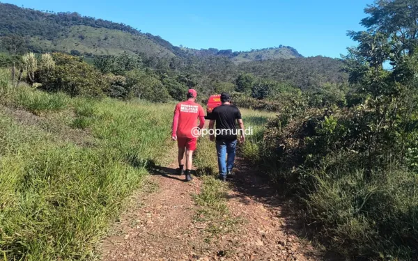 Duas pessoas caminham por um caminho em meio à vegetação e a uma colina. A composição da cena mostra um caminho de terra que serpenteia por uma área de vegetação, com duas figuras humanas caminhando na direção oposta. A posição das pessoas sugere um ponto de vista de trás para a frente. O foco da imagem está principalmente nas figuras, com o terreno circundante desfocado. Os elementos são colocados para destacar as pessoas caminhando no caminho. As duas pessoas são adultas e parecem caminhar em direção ao fundo da imagem. As cores dos seus trajes são vermelhas e pretas, contrastando com o verde do fundo. Os detalhes específicos da vestimenta são mais visíveis quanto aos seus tecidos e estilo. O cenário é composto por um caminho de terra batida através de um terreno aberto com vegetação baixa e uma colina ao fundo. A iluminação é natural e clara, indicando um dia ensolarado. A atmosfera é tranquila e sem emoções fortes.