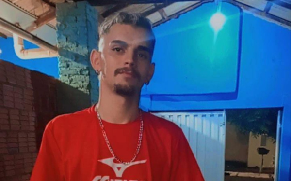 Jovem com cabelo curto e claro, barba e bigode ralos, usando brinco na orelha esquerda. Ele veste uma camiseta vermelha com estampa branca e corrente prateada. A foto foi tirada à noite, sob uma estrutura coberta, com uma parede azul ao fundo iluminada por uma lâmpada.