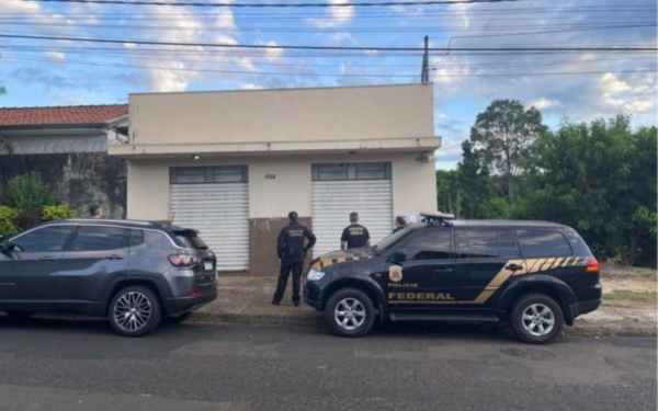 Imagem de uma operação da Polícia Federal em frente a um imóvel de fachada simples, com portas de metal fechadas. Três agentes, vestidos com uniformes pretos e coletes, estão posicionados diante do local. Duas viaturas estão estacionadas na rua: uma SUV preta identificada como da Polícia Federal e um veículo cinza ao lado. O cenário inclui um céu parcialmente nublado, árvores ao fundo e fiação elétrica sobre a via.