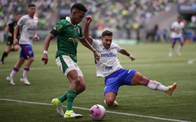 A composição da cena mostra dois jogadores de futebol em uma ação de disputa intensa. O jogador de camisa verde está em movimento, enquanto o outro jogador de camisa azul está em uma posição de queda. O jogador verde parece estar em uma posição de controle da bola, enquanto o jogador azul está se posicionando para interceptar ou disputar a bola. O foco está principalmente nos dois jogadores principais envolvidos na disputa.