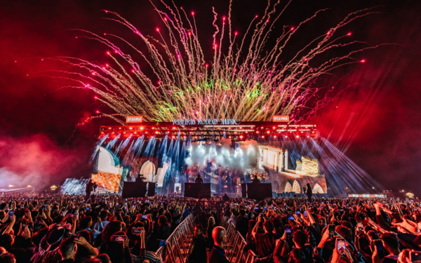 Palco iluminado do Ribeirão Rodeo Music 2025 à noite, com fogos de artifício coloridos no céu. Uma multidão de pessoas lota a arena, muitas segurando celulares para registrar o momento. No palco, luzes vibrantes e imagens de temática sertaneja completam o cenário da festa