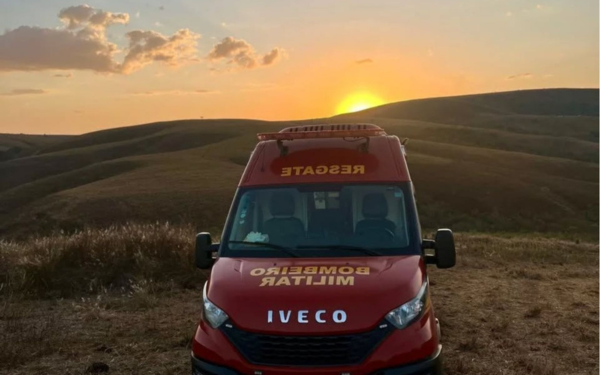 A imagem mostra uma ambulância vermelha de resgate do Corpo de Bombeiros Militar, estacionada em uma área de campo aberto. No horizonte, o sol está se pondo atrás das colinas, iluminando o cenário com luz alaranjada. O céu está parcialmente coberto por nuvens.