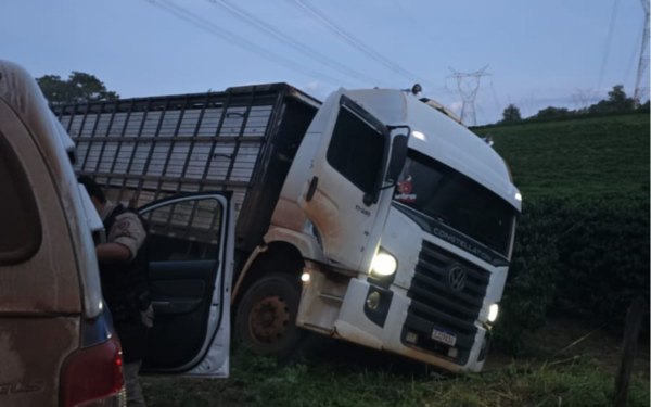 Um caminhão branco está inclinado em um terreno acidentado, possivelmente fora da estrada. Há um carro ao lado dele, e parece que existe a presença de autoridades, com pessoas de uniforme.