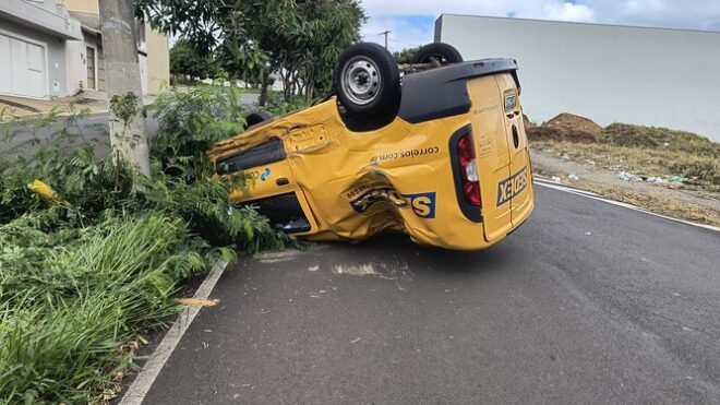 Veículo amarelo dos Correios capotado, com as rodas para cima, em uma rua asfaltada. O carro está tombado ao lado de um poste e parcialmente encoberto por vegetação. O acidente ocorreu próximo a uma calçada e uma área com mato e entulho. O tempo está nublado.