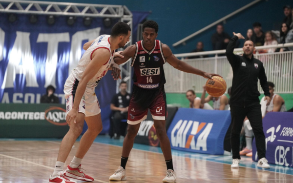 Em uma quadra de basquete, dois jogadores disputam uma jogada. À esquerda, um atleta do Pato Basquete, com uniforme branco e detalhes em vermelho e azul, está em posição defensiva, com os joelhos flexionados. À direita, Georginho, do Franca Basquete, usa uniforme vinho e preto com o número 14. Ele segura a bola com a mão direita, pronto para iniciar o drible, encarando o marcador com foco. Ao fundo, o técnico da equipe francana, de roupa preta, gesticula orientando o time. Painéis de patrocinadores e uma grande faixa azul com o nome “PATO” compõem o ambiente.