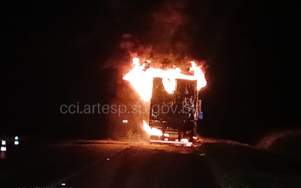 Foto tirada à noite mostra a traseira de uma carreta em chamas no meio da rodovia. As labaredas intensas consomem a parte superior e lateral do veículo, iluminando a escuridão ao redor. Cones de sinalização são visíveis à esquerda da pista, indicando interdição parcial. A imagem tem a marca d'água “cci.artesp.sp.gov.br”.