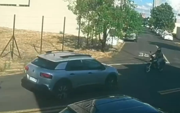 Imagem de câmera de segurança mostra o momento exato de uma colisão entre um carro e uma motocicleta em uma rua de bairro residencial. Um veículo Citroen prata faz uma conversão à esquerda, cruzando a pista, enquanto uma motociclista, vindo em sentido contrário, está prestes a colidir com a lateral dianteira do carro. A rua é asfaltada, com árvores e calçadas ao fundo. O impacto é iminente