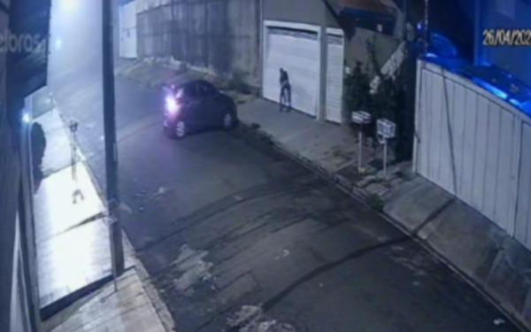 Imagem de uma rua residencial durante a noite. À direita, um homem aparece forçando o portão de uma casa, enquanto um carro de cor escura, com os faróis acesos, está parado em frente à calçada. A rua está deserta e iluminada por postes de luz. Câmeras de segurança registram a ação. No canto superior direito da imagem, aparece a data "26/04/2025"
