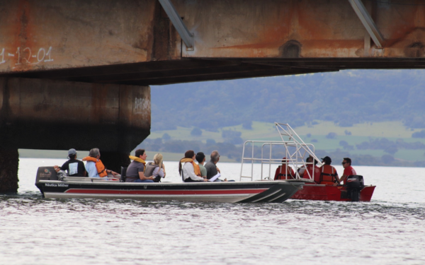Duas embarcações com equipes técnicas navegam sob a ponte que liga Rifaina (SP) a Sacramento (MG), durante vistoria na estrutura. As pessoas usam coletes salva-vidas e observam a parte inferior da ponte
