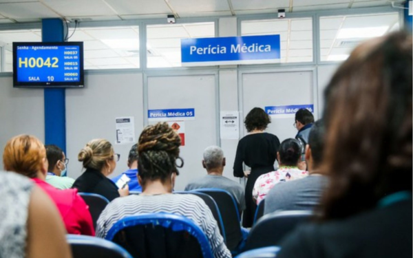Sala de espera de uma unidade de perícia médica do INSS. Várias pessoas estão sentadas em cadeiras azuis, de costas para a câmera, aguardando atendimento. Algumas usam máscaras. À frente, há duas portas com placas indicando "Perícia Médica 05" e "Perícia Médica 06". Acima das portas, uma faixa azul com a inscrição “Perícia Médica”. À esquerda da imagem, uma tela eletrônica exibe senhas e salas de atendimento
