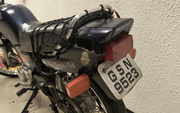 Imagem mostra a parte traseira de uma motocicleta encostada em uma parede de textura rústica. A moto está visivelmente desgastada e tem uma placa artesanal com os caracteres "GSN 9523", feitos de forma irregular e desalinhada. A placa é branca com letras pretas, sem padrão oficial. Acima da lanterna traseira, há um bagageiro com elásticos entrelaçados. Do lado esquerdo da rabeta, há um adesivo com a frase “Sou louco por Rock” e um rosto estilizado com óculos escuros