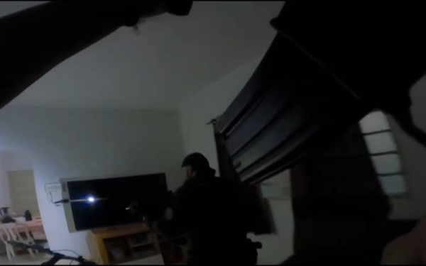 Imagem mostra o interior de uma residência durante o cumprimento de um mandado pela Polícia Civil. Em primeiro plano, vê-se parcialmente o braço de um policial com uma arma apontada. Ao fundo, outro agente do GOE (Grupo de Operações Especiais) caminha em direção à sala, iluminando o ambiente com uma lanterna acoplada à arma. O local parece tranquilo, com uma TV ligada e uma mesa de jantar ao fundo. A cena é registrada sob baixa luminosidade, sugerindo uma ação durante a madrugada.