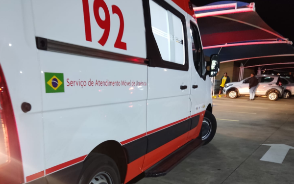 Ambulância branca e vermelha do SAMU (Serviço de Atendimento Móvel de Urgência), com o número 192 em destaque, está estacionada em um pátio iluminado à noite. À frente do veículo, há algumas pessoas próximas a um carro prata sob uma cobertura de lona. O cenário sugere uma ocorrência em andamento, possivelmente em um estacionamento de supermercado.