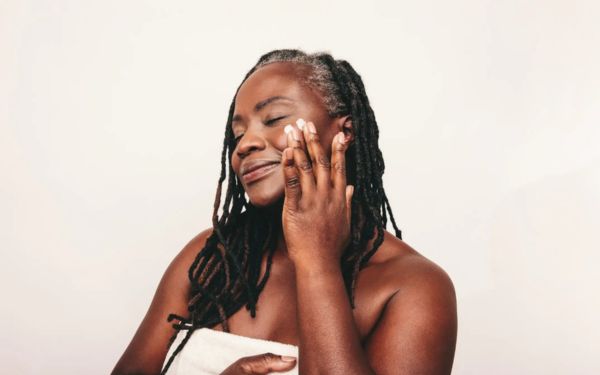A imagem mostra uma mulher negra, de idade mais madura, passando um creme no rosto, mosgtrando cuidados com a pele