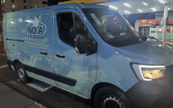 Fotografia noturna de uma van funerária azul-clara da empresa "Nova Franca Assistência Familiar", estacionada em uma vaga reservada em frente a um posto de combustíveis da rede JetOil. O veículo tem adesivos com nuvens e o slogan "Sempre valorizando a vida". No banco do motorista, há um homem com camiseta verde, visível através da janela aberta. O entorno está iluminado por luzes artificiais.