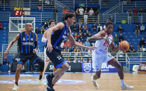 Imagem de uma partida de basquete entre Franca e Pinheiros no ginásio Pedrocão. O jogador do Franca Basquete, com uniforme branco e número 14, está em movimento com a bola nas mãos, tentando escapar da marcação. Dois atletas do Pinheiros, vestindo uniformes pretos com listras azuis, tentam bloqueá-lo. Ao fundo, há torcedores nas arquibancadas acompanhando a partida.