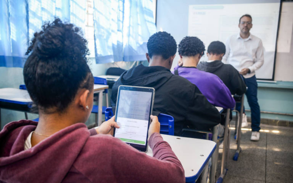 Em uma sala de aula iluminada pela luz azulada que entra pelas janelas, alunos estão sentados em carteiras escolares, de costas para a câmera. Uma aluna, em destaque à esquerda, segura um tablet onde aparece a plataforma educacional TarefaSP. Ela veste um moletom roxo escuro e tem o cabelo preso. Ao fundo, o professor está em pé diante da lousa, orientando a turma.