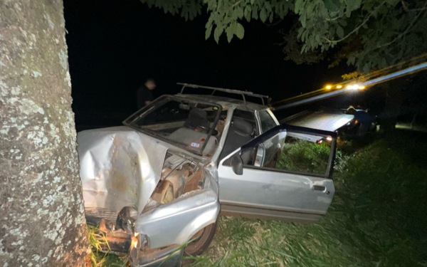 Imagem noturna mostra um carro prata modelo Escort com a parte dianteira totalmente destruída após colidir com uma árvore de grande porte à margem de uma estrada. A porta do motorista está aberta, e o veículo está em uma área de vegetação. Ao fundo, há um guincho com luzes acesas e uma pessoa desfocada