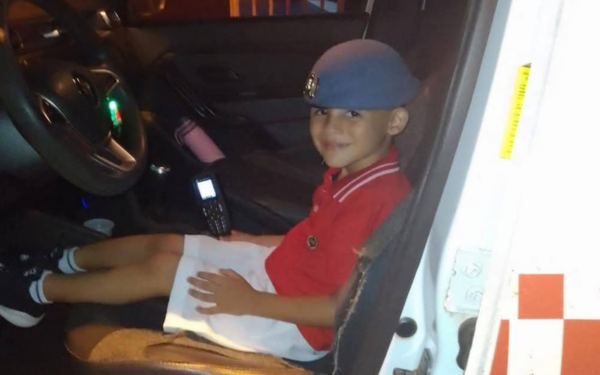 Menino de aproximadamente seis anos está sentado no banco do motorista de um carro, sorrindo. Ele veste camiseta vermelha, bermuda branca, tênis preto com meias brancas e um boné azul com brasão. À sua frente, no painel do veículo, há um rádio comunicador.