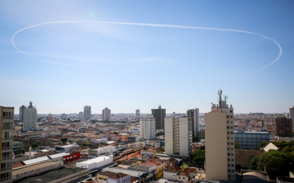 A imagem mostra a cidade de Franca (SP) vista de um ponto alto, com destaque para a região central repleta de prédios residenciais e comerciais. No céu limpo e azul, uma trilha branca formada por um avião desenha um grande círculo quase perfeito, chamando a atenção pela sua dimensão e simetria. A cena une o cotidiano urbano ao inusitado espetáculo aéreo