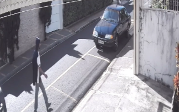 Imagem de uma câmera de segurança capturada durante o dia em uma rua residencial de Franca. À esquerda, um homem anda pela calçada vestindo calça clara, camiseta branca com listras escuras e capuz cobrindo a cabeça. À direita da imagem, está estacionado um carro Monza azul-escuro, com o capô refletindo a luz do sol. O local é cercado por muros brancos e vegetação
