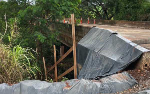 Imagem mostra uma ponte antiga interditada sobre uma área de mata, com sinais visíveis de comprometimento estrutural. Parte da lateral da ponte está coberta com uma lona preta, sustentada por escoras de madeira. No topo da estrutura, cones de sinalização laranja estão posicionados para impedir a passagem de veículos. A vegetação ao redor é densa, com árvores e mato alto próximos à construção.