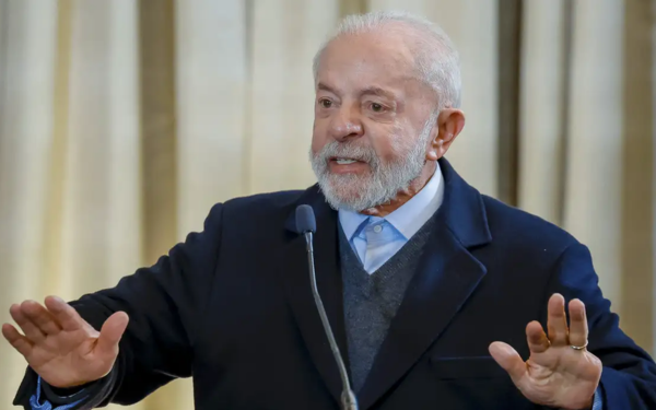 O presidente Luiz Inácio Lula da Silva aparece em pé, atrás de um microfone, fazendo um gesto com as mãos levantadas à frente do corpo, como se estivesse enfatizando algo ou pedindo calma. Ele usa paletó escuro, camisa azul clara e suéter cinza por baixo. Ao fundo, cortinas bege desfocadas completam o cenário