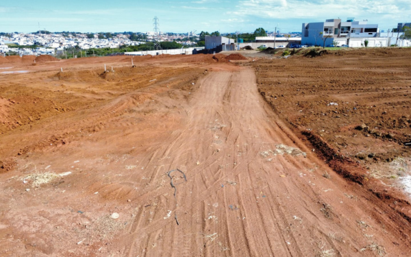 Imagem de uma área em obras, com chão de terra avermelhada e marcas de pneus. No centro, uma via de terra batida segue em direção ao fundo da imagem, onde há algumas edificações e muros brancos. Do lado direito, terrenos nivelados indicam terraplenagem recente. Ao fundo, é possível ver casas, prédios e uma torre de energia ao longe, sob um céu azul com nuvens dispersas.