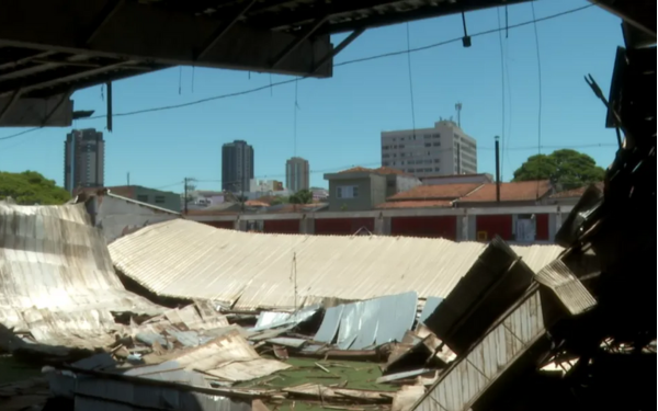 Imagem mostra o interior do ginásio do Clube dos Bagres, em Franca (SP), com o teto completamente desabado. Estruturas metálicas retorcidas e pedaços de telhado estão espalhados pelo chão. Ao fundo, é possível ver prédios residenciais e comerciais da cidade, contrastando com os destroços no primeiro plano. A luz do dia ilumina a cena, evidenciando o estado de abandono da estrutura.