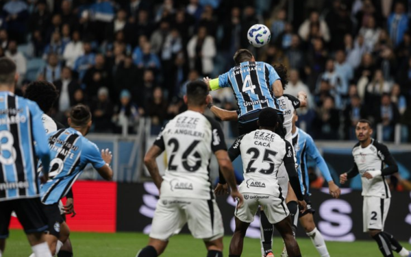 Jogadores de Grêmio e Corinthians disputam a bola pelo alto durante partida na Arena do Grêmio. Um atleta do Grêmio, usando o uniforme azul celeste e preto, sobe mais alto que os adversários e cabeceia a bola, enquanto outros jogadores dos dois times observam a jogada. A arquibancada ao fundo está cheia de torcedores, muitos com roupas nas cores do Grêmio.
