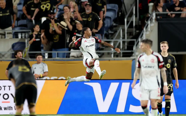 Jogador do Flamengo, vestindo uniforme branco com detalhes vermelhos, salta no ar comemorando um gol com o punho cerrado e expressão de euforia. Ao fundo, torcedores do Los Angeles FC, vestidos de preto e dourado, acompanham a cena. Outros atletas dos dois times estão no campo, alguns ainda reagindo à jogada.