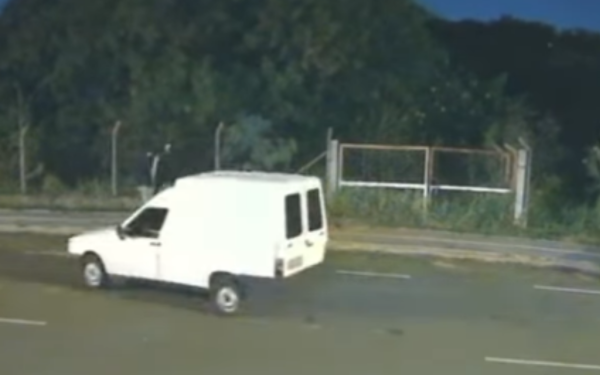 Imagem de uma rua durante a noite. Uma Fiorino branca aparece em movimento na pista, indo da esquerda para a direita. Ao fundo, há uma área de mata fechada e escura, cercada por uma cerca de arame e portões metálicos. A cena foi registrada por uma câmera de segurança.