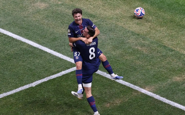 Dois jogadores do Paris Saint-Germain comemoram um gol com empolgação dentro da grande área. Um deles está no ar, abraçado ao companheiro, enquanto sorri largamente. Ambos vestem o uniforme azul escuro do PSG. A bola aparece no gramado, rolando ao fundo. A cena transmite alegria e euforia do momento do gol.