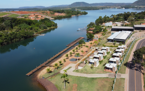 A imagem mostra uma vista aérea da margem da Represa de Jaguara, em Sacramento (MG), onde está localizado um moderno complexo turístico. À esquerda, a represa de águas calmas se estende entre vegetações e encostas. Na margem, vê-se um longo píer de madeira sobre a água, palmeiras alinhadas em uma faixa de areia artificial e uma estrutura de lazer com pequenos chalés ou bangalôs brancos organizados em fileiras. Ao lado, uma construção ampla com cobertura metálica parece ser o restaurante ou clube principal. Ao fundo, a paisagem é cercada por morros e vegetação. Uma estrada asfaltada contorna a área do complexo.