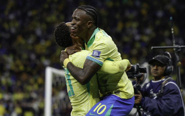 Imagem mostra o atacante Vinicius Júnior comemorando seu gol com os braços erguidos, ainda dentro da área adversária. Ele veste o uniforme tradicional da seleção brasileira, com camisa amarela e calção azul. Ao fundo, torcedores nas arquibancadas celebram e aplaudem, enquanto companheiros de equipe correm em sua direção para comemorar o gol.