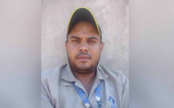 Homem de pele parda, barba rala e cabelo curto, usando boné preto com detalhe amarelo na aba. Ele veste uma camisa de uniforme bege com crachá pendurado no pescoço e está posicionado de frente para a câmera, com expressão séria. Ao fundo, uma parede lisa de concreto claro.