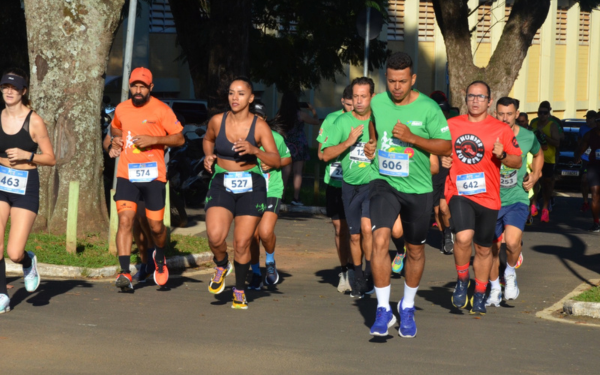 Grupo de corredores participa de uma prova de rua durante o dia. Eles estão em movimento, vestindo roupas esportivas e números de identificação presos na frente das camisetas. Há homens e mulheres entre os participantes, com expressões concentradas. O ambiente é arborizado e a corrida acontece em uma via asfaltada, com árvores e um prédio ao fundo.