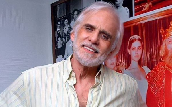 Homem idoso de cabelos e barba brancos sorri levemente, vestindo uma camisa listrada em tons claros. Ele está em pé, em frente a uma parede decorada com fotos antigas em preto e branco e coloridas, que remetem ao teatro e à televisão.