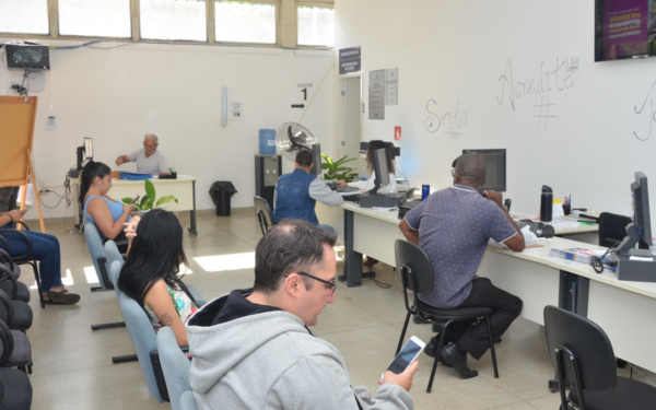 Sala de atendimento do programa Emprega Franca. À esquerda, há cadeiras com pessoas aguardando, algumas mexendo no celular. À direita, atendentes estão sentados em frente a computadores, conversando com candidatos. Ao fundo, outro servidor trabalha em uma mesa isolada. A sala é iluminada, com paredes brancas e frases motivacionais escritas, como "Sorria" e "Acredite"