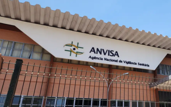 Fachada do prédio da Anvisa (Agência Nacional de Vigilância Sanitária), com uma grande placa branca exibindo o logotipo da instituição — formado por elementos gráficos verdes, amarelos e pretos ao lado do nome “ANVISA”. O edifício tem parede de tijolos à vista, janelas envidraçadas e cobertura metálica em estilo ondulado. Na frente do prédio, há uma grade de proteção preta.