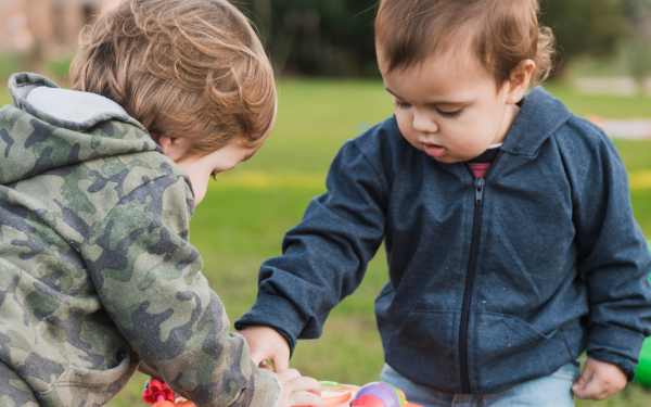 Dois meninos pequenos, com cerca de dois anos de idade, estão ao ar livre, concentrados em uma brincadeira. Um veste moletom azul escuro e o outro usa uma jaqueta camuflada. Ambos estão debruçados sobre uma mesinha com brinquedos coloridos. O ambiente é gramado, com fundo desfocado, indicando um espaço amplo e tranquilo.