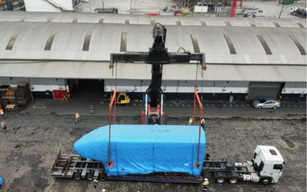 Imagem aérea mostra um grande galpão industrial com um caminhão estacionado em frente. No centro da foto, um guindaste suspende uma parte de trem, coberta com uma lona azul, que está sendo cuidadosamente posicionada sobre a carroceria do caminhão. Algumas pessoas com coletes refletores acompanham a operação no chão, indicando se tratar de uma movimentação logística importante. O local tem piso de paralelepípedo e aparência de área portuária ou industrial.