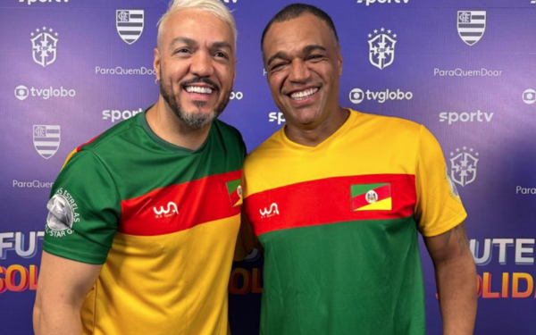 Na imagem, o cantor Belo e o ex-jogador Denilson aparecem lado a lado, sorrindo. Belo está com os cabelos loiros, usa camiseta branca e colar prateado. Denilson veste uma camisa preta e também sorri. Ambos estão próximos, em um ambiente iluminado, transmitindo um clima descontraído e amigável.