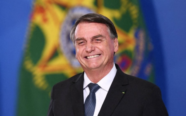 O ex-presidente Jair Bolsonaro aparece sorrindo, vestindo terno preto, camisa branca e gravata azul. Ao fundo, desfocada, está a bandeira do Brasil.
