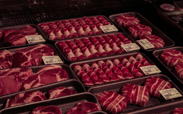 Bandejas de carne bovina estão expostas em um balcão refrigerado de supermercado. As peças estão organizadas em fileiras, embaladas em plástico transparente e com etiquetas de preço visíveis. Há cortes variados, incluindo bifes, carnes enroladas com bacon e peças inteiras, todas com coloração vermelho-viva, indicando frescor.