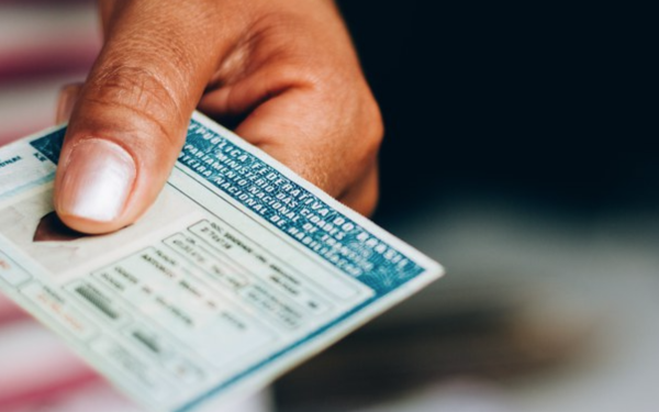 Imagem mostra uma mão segurando uma Carteira Nacional de Habilitação (CNH) azul, tradicional no Brasil. A mão aparece em destaque, com unhas curtas e levemente polidas. O fundo está desfocado, direcionando a atenção para o documento.