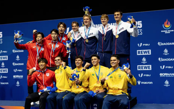 Três equipes de nadadores posam no pódio após a prova de revezamento 4x100 metros livre nos Jogos Mundiais Universitários. No topo, os atletas dos Estados Unidos comemoram com medalhas de ouro e mascotes na mão. Ao lado, os japoneses, vestidos de vermelho, exibem as medalhas de prata. Abaixo, sentados, os brasileiros com agasalhos amarelos e verdes, seguram as medalhas de bronze e os mascotes do evento, sorrindo para a foto.