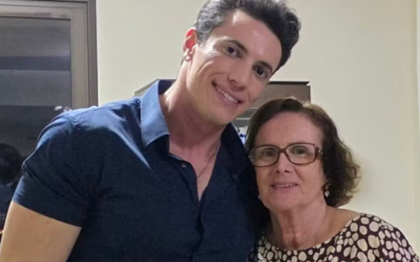 Um homem jovem, de pele clara, cabelo escuro e penteado para o lado, está abraçado a uma senhora mais velha, também de pele clara, com óculos e cabelos castanhos curtos. Ele veste uma camisa azul escura e sorri levemente para a câmera. A senhora, que está com uma blusa estampada de bolinhas claras, também sorri com expressão serena. Os dois estão em um ambiente interno com paredes claras.
